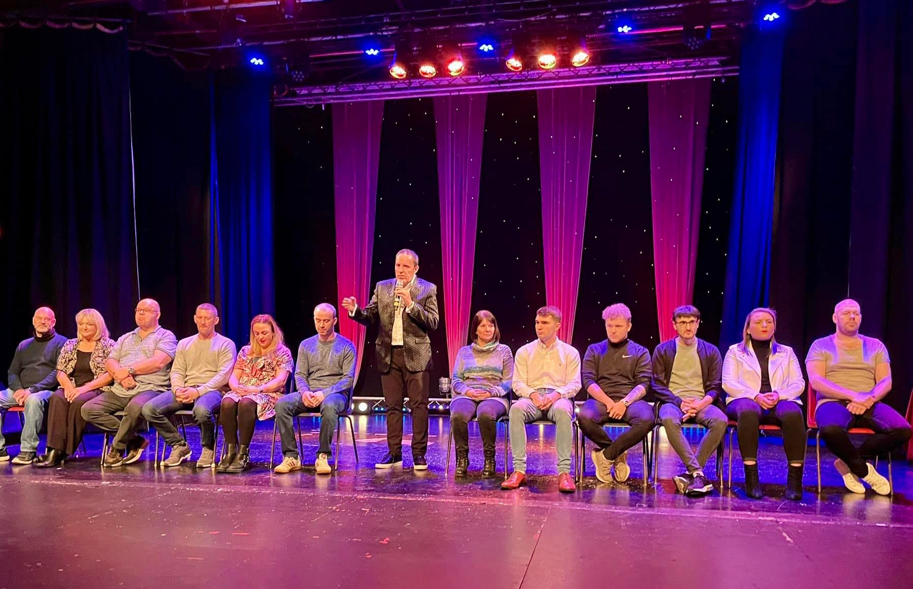 Volunteers relaxed in chairs on stage at a comedy hypnosis show while one stands laughing and the audience applauds